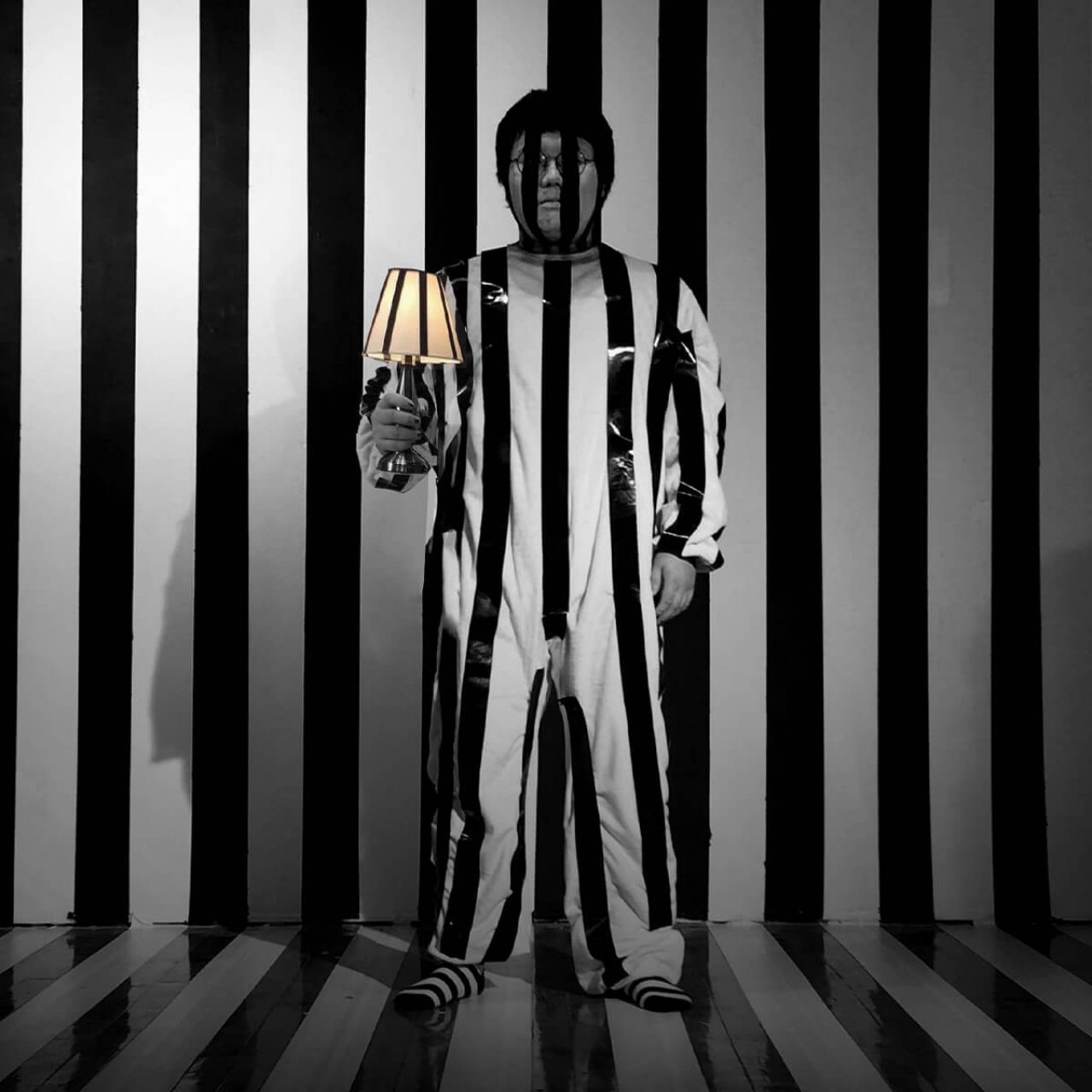 松林李站在lamp and shade in his hand against a black and white vertically striped backdrop. The lamp and suit he wears is also striped.
